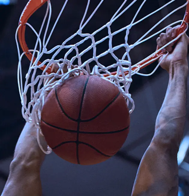 A basketball is being dunked through a hoop from below, with hands gripping the rim. The net is partially visible, and the background is dark, with focus on the action.