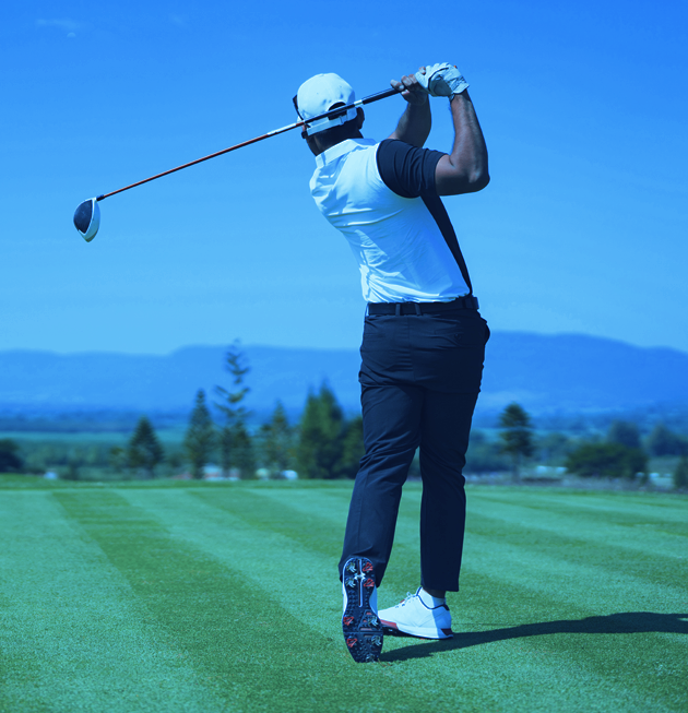 A golfer, symbolizing wealth and sports, wears a white cap, white shirt, and black pants as he finishes a swing on a lush green course with trees and mountains in the background beneath a clear blue sky.